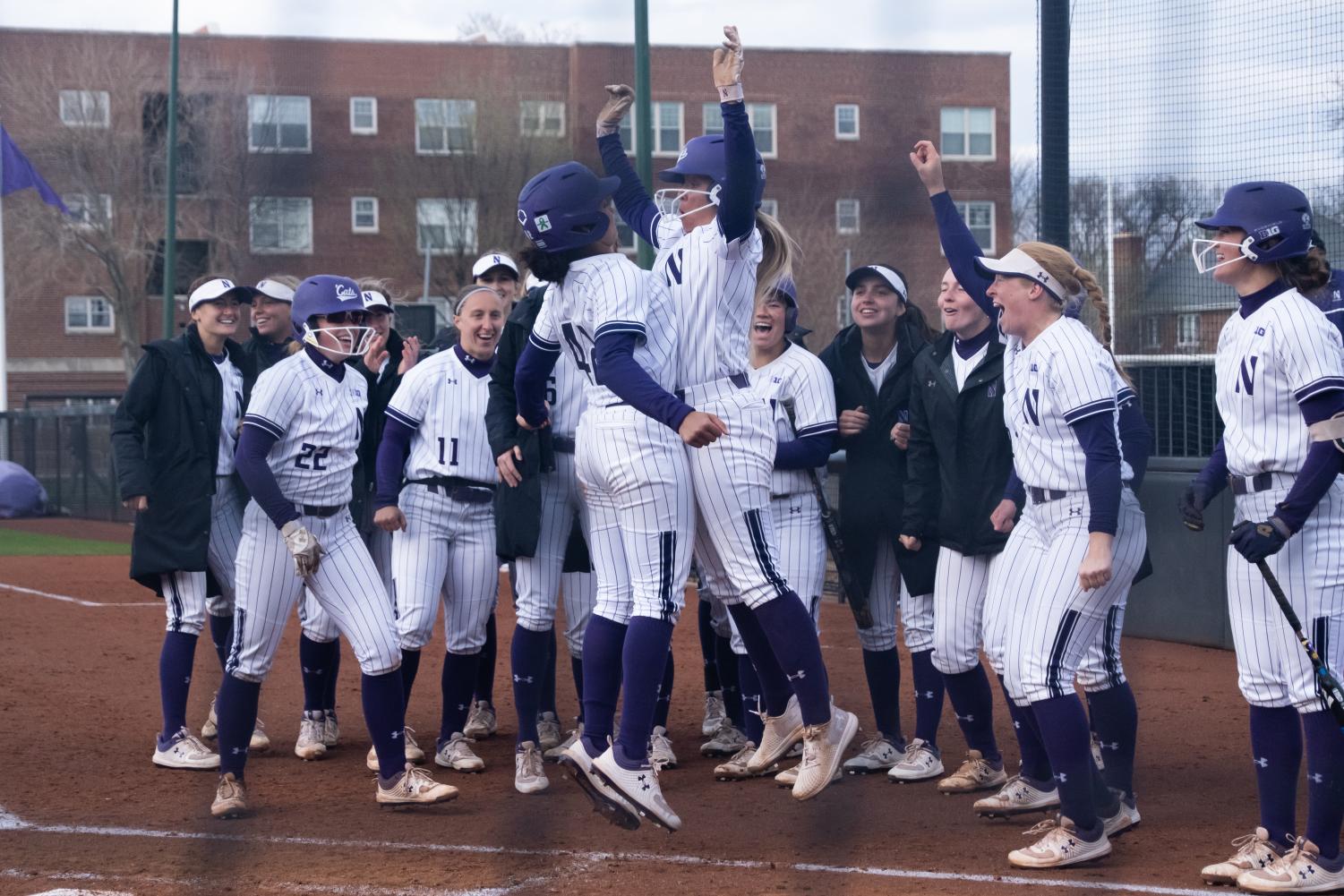 A+group+of+softball+players+in+white+uniforms+with+purple+stripes+are+jumping+and+celebrating+together.