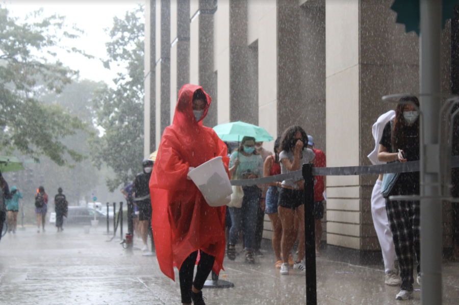 A+person+in+a+red+poncho+holds+a+white+bucket+while+students+stand+in+line+against+a+tan+building+in+the+rain.