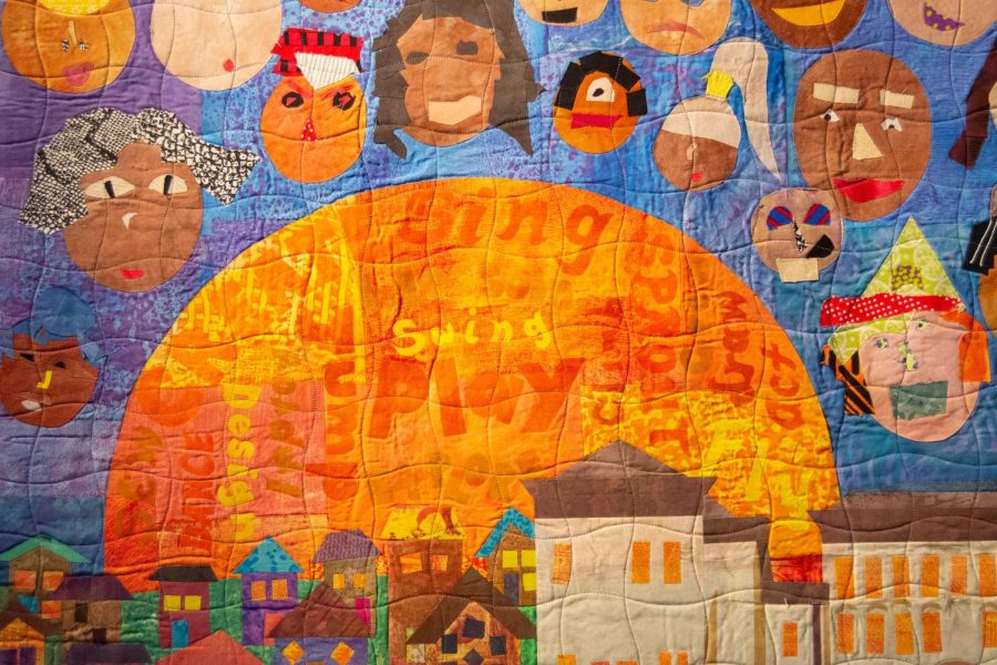 A close up photo of Maggie Weiss’ quilt, showing quilted portraits of faces and a large sun featuring words such as “play” and “sing.”