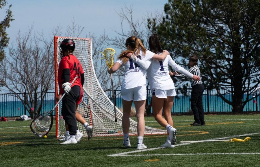 A player wearing a white and purple jersey with the number ‘18’ on it embraces another player wearing a white and purple jersey with the number ‘1’ on it. The two walk toward a lacrosse goal, where a goalie wearing red stands with a ball in the net.