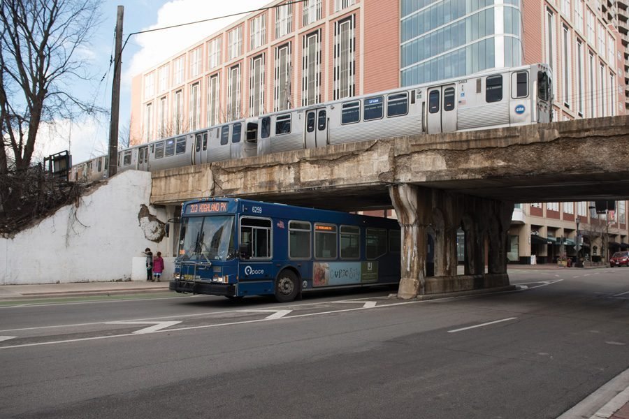 A blue bus passes under a gray overpass while a train goes by above.