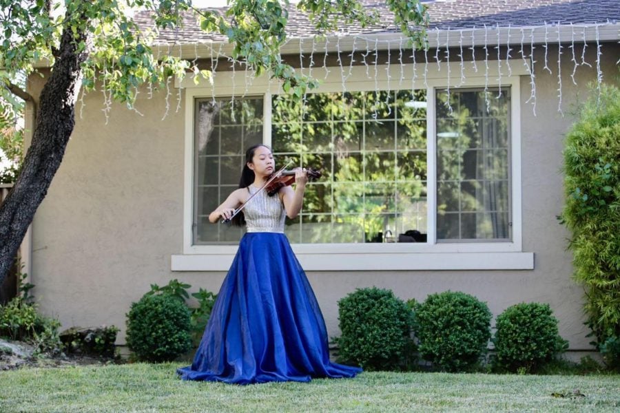 A+woman+playing+the+violin+wearing+a+blue+and+silver+gown.+The+background+has+a+tan+building%2C+green+bushes%2C+and+grass.
