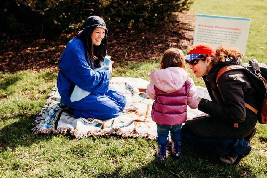 A woman in costume acts out the story of Passover at JUF’s Young Families Program Passover celebration at Morton Arboretum. She sits on a blanket on the grass smiling at a little girl. The girl’s mom crouches next to her and speaks to her.