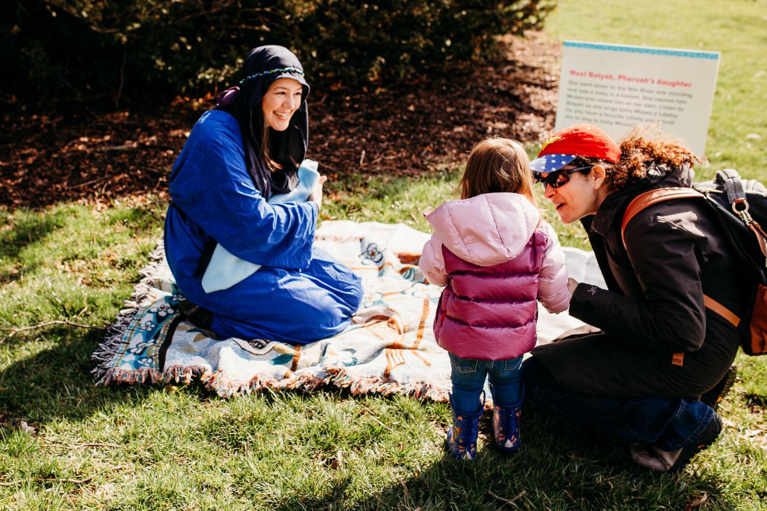 A+woman+in+costume+acts+out+the+story+of+Passover+at+JUF%E2%80%99s+Young+Families+Program+Passover+celebration+at+Morton+Arboretum.+She+sits+on+a+blanket+on+the+grass+smiling+at+a+little+girl.+The+girl%E2%80%99s+mom+crouches+next+to+her+and+speaks+to+her.