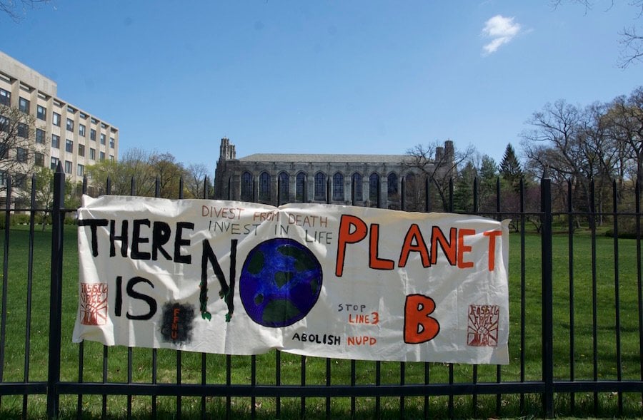 A white banner reads “There is no Planet B” and hangs from a black fence in front of a green lawn and gray library building.