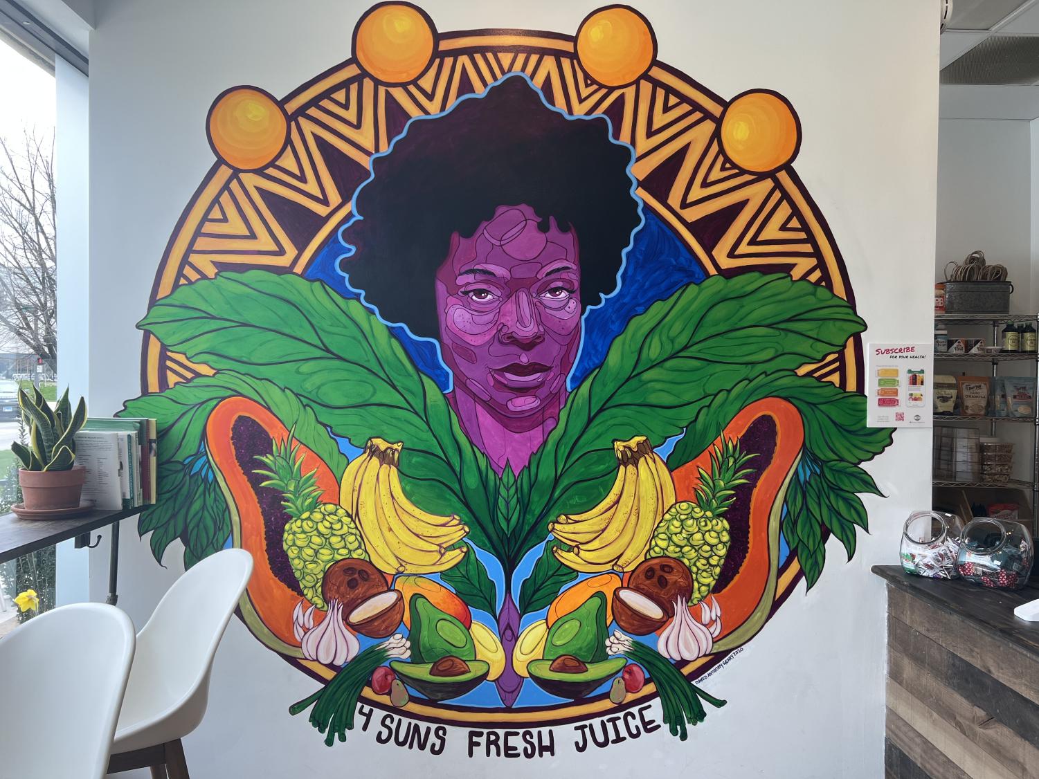 A+mural+of+a+Black+woman+surrounded+by+green+plants+and+various+colorful+fruits+on+a+white+wall.+She+is+surrounded+by+a+sun+design+and+the+words+%E2%80%9C4+Suns+Fresh+Juice%E2%80%9D+line+the+bottom+of+the+mural.