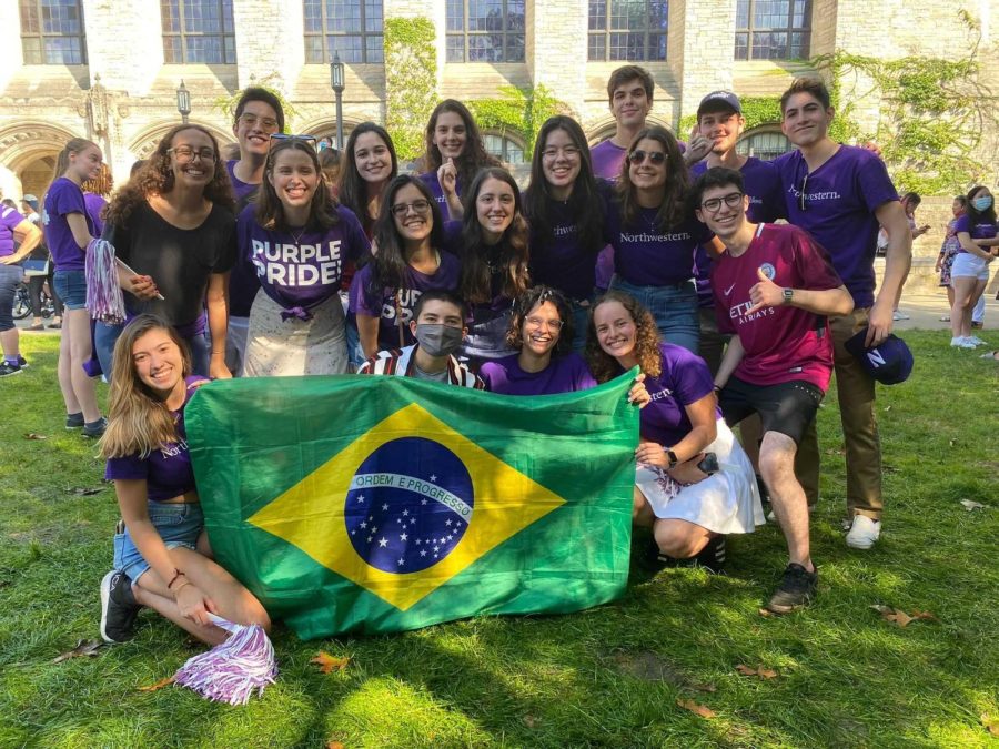 A+group+of+people+pose+outdoors+with+a+Brazilian+flag.