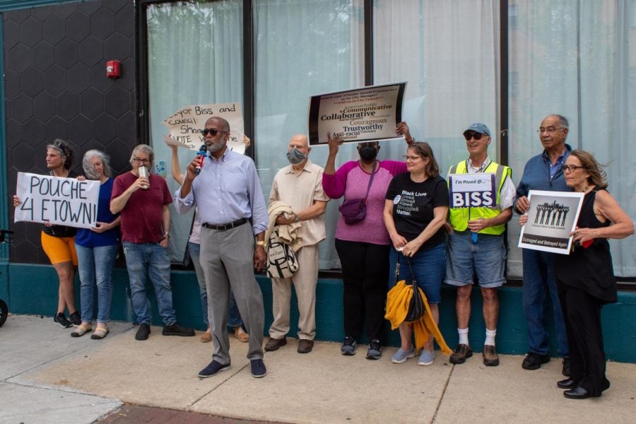 Several protesters stand on a sidewalk outside a building, holding signs reading “Poche 4 ETown” and “I’m Pissed at Biss.” A man holding a microphone stands in the middle, speaking to onlookers.