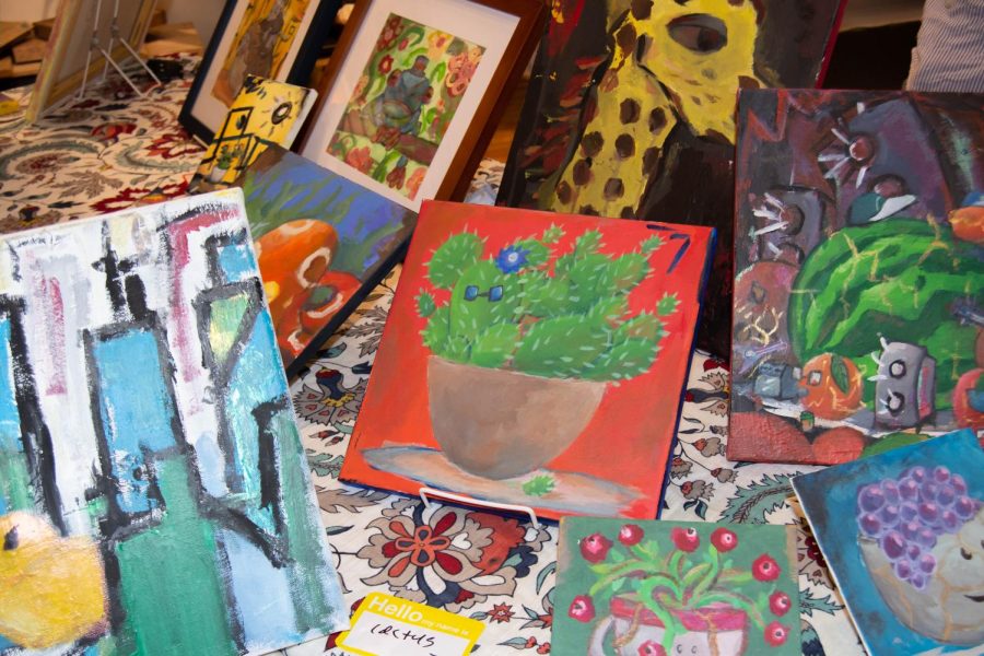 A painting of a green cactus with glasses and a blue flower in a clay bowl on a red background sits in the center of a table. It is surrounded by other paintings including a yellow giraffe, abstract skyline and fruit with faces.