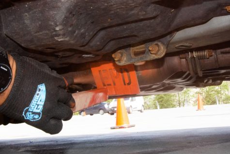 The underside of a car is shown. A gloved hand fills in an “EPD” stencil with bright orange spray paint on a catalytic converter.