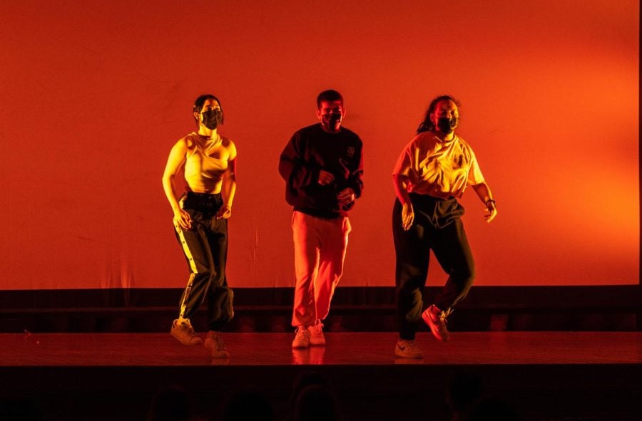 Three dancers stand on stage with red lighting.