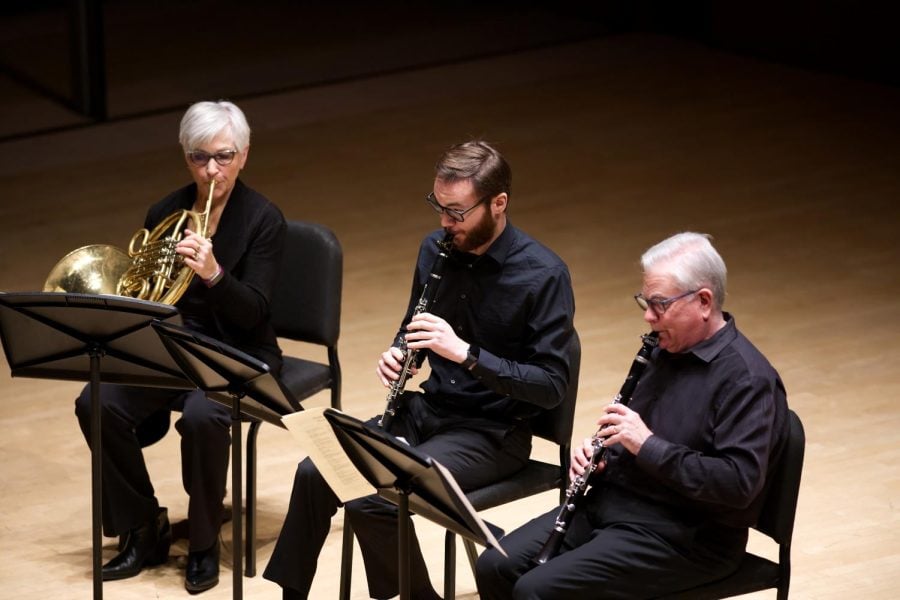 Three people play instruments (left to right: french horn, clarinet, clarinet) on a wooden stage. They wear all black and sit facing three music stands.