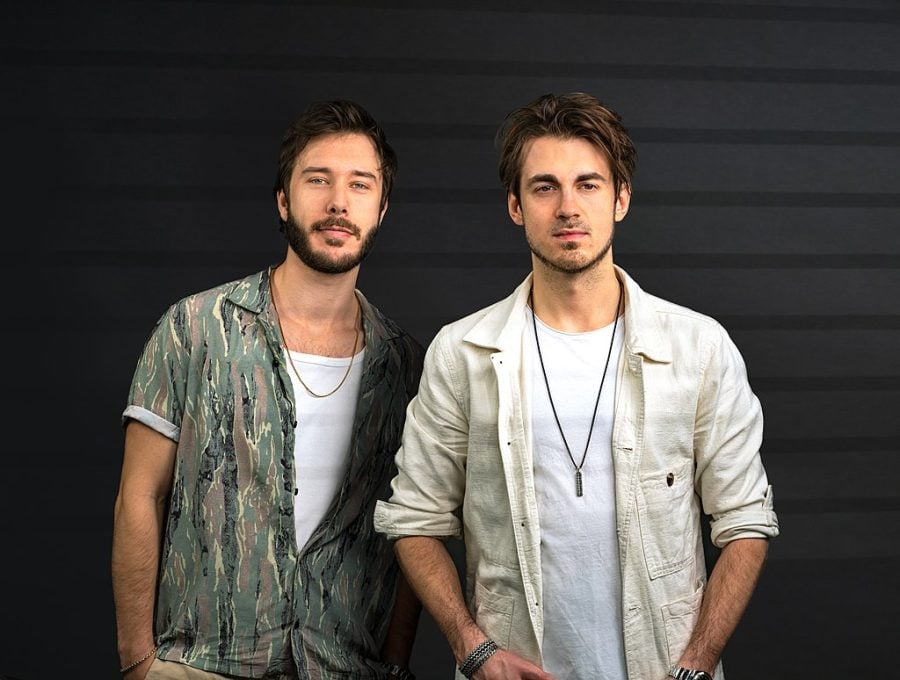 Two people in open button downs and white tank tops stand in front of a gray wall.