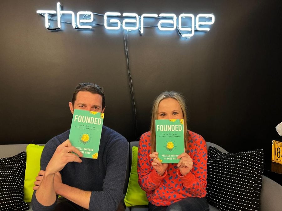 Mike Raab and Melissa Kaufman sitting on a gray couch, holding their new book in front of a neon sign reading, The Garage.