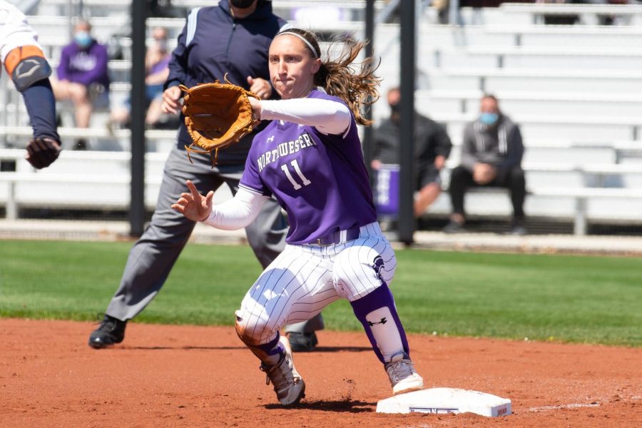 Softball player wearing purple uniform holds out glove on field.