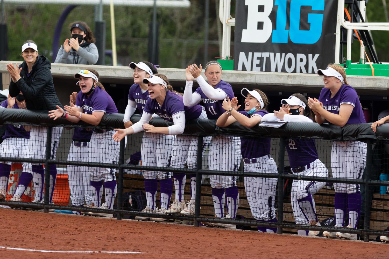 Seven+girls+in+purple+jerseys+and+white+pants+cheer+and+smile+from+a+softball+dugout.