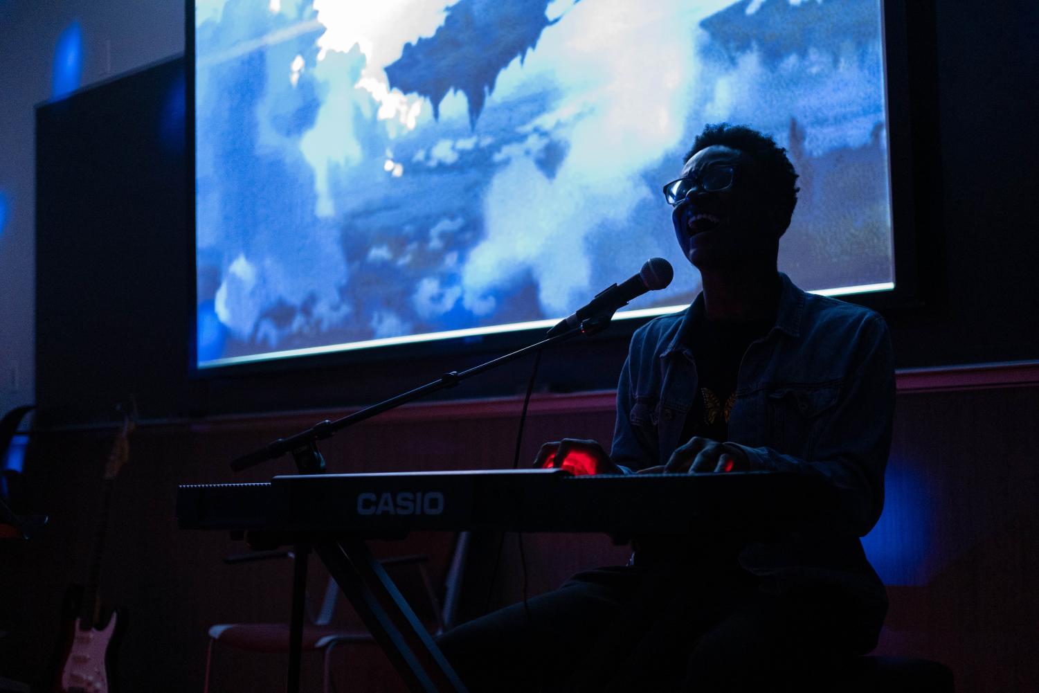  A performer plays a keyboard and sings into a microphone in a dark room with a blue background.