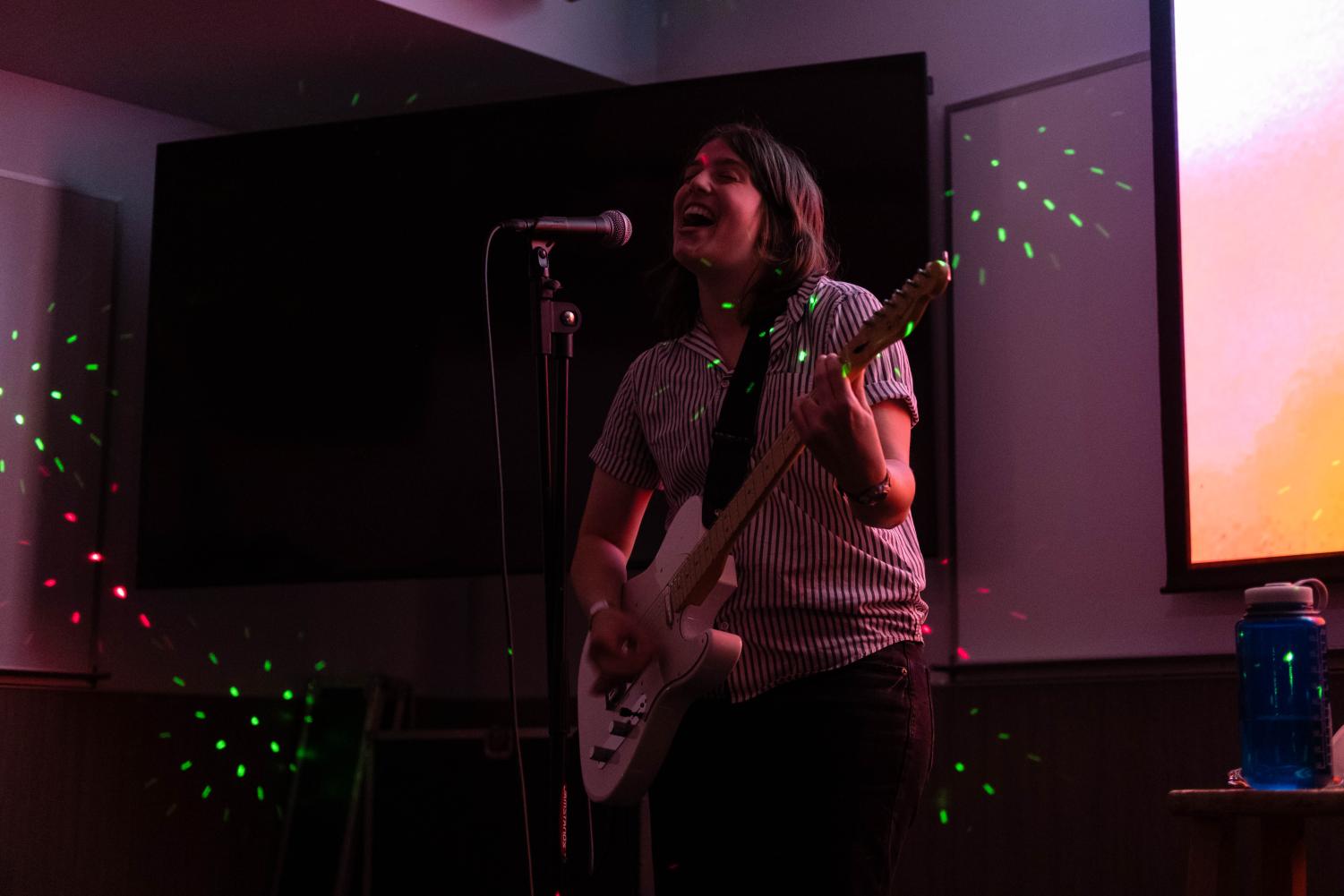 A performer plays an electric guitar and sings into a microphone in a dimly lit room. They wear a striped shirt and green dotted lights shine around them.