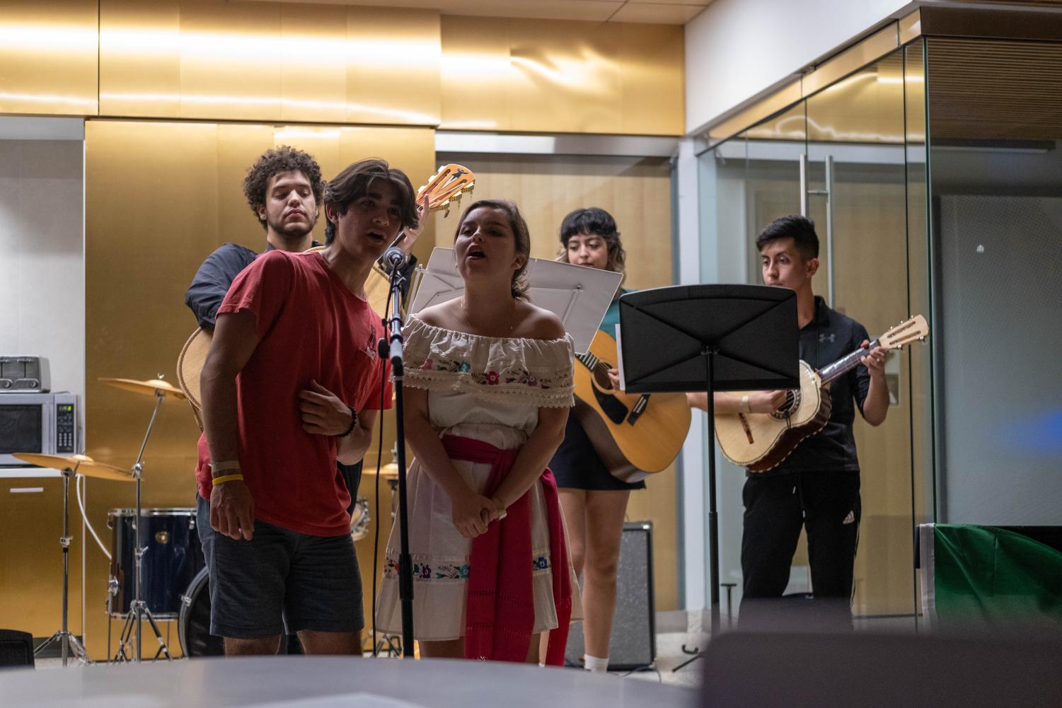 A student mariachi group performs. Two performers sing into a microphone as three others play instruments behind them.