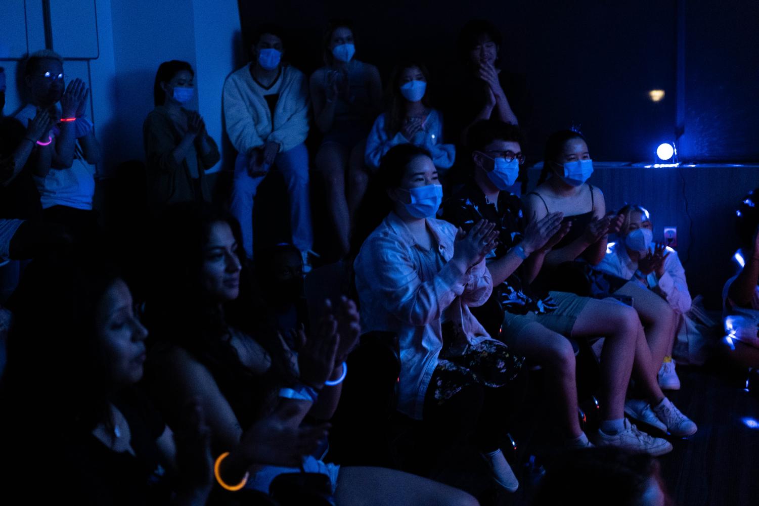 A crowd of people clap in a room with dim blue lighting.