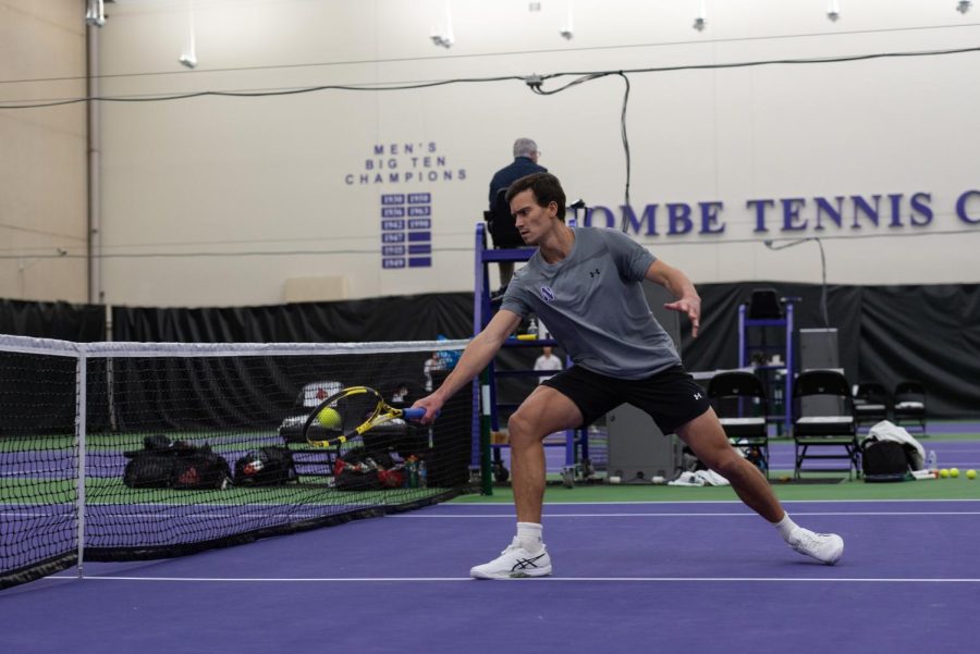 Tennis player in gray shirt, white shoes and black shorts meets ball with tennis racquet near the net on purple and green court.