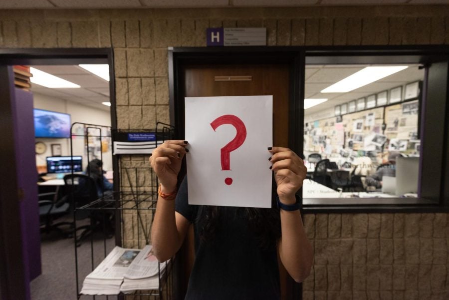A person holds up a white piece of paper with a big red question mark in front of their face. The Daily Northwestern’s newsroom is in the background.