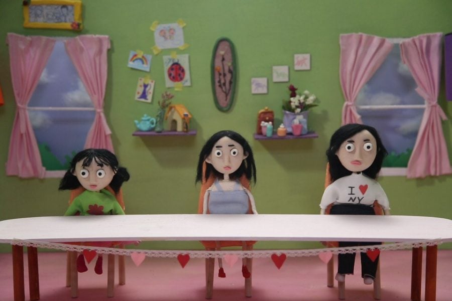 Three dolls in a stop-motion still are positioned at a table facing the camera. From right to left: The first is a child with short black pigtails and a green sweater, the second has mid-length black hair and the third is an adult with long black hair. The background is a green wall with windows that have pink curtains.