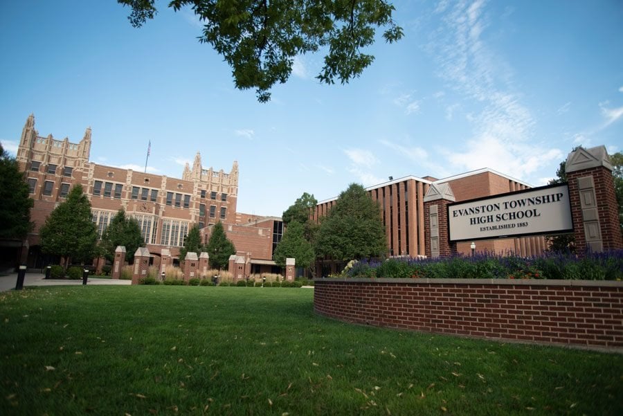 Green grass in the foreground and large red-brick school buildings in the background. A sign in the front reads “Evanston Township High School.”