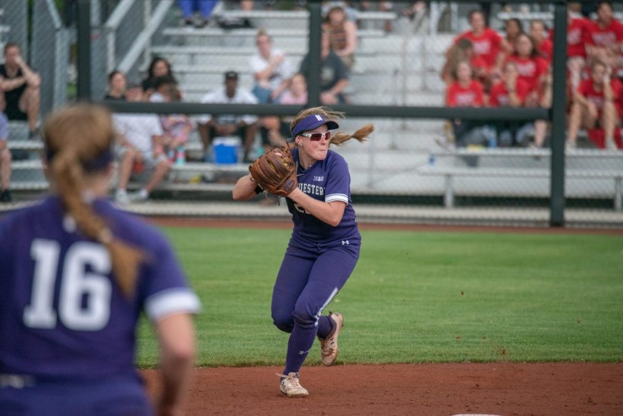Softball+player+wearing+purple+uniform+and+sunglasses+holds+ball+in+glove+on+a+dirt+basepath.