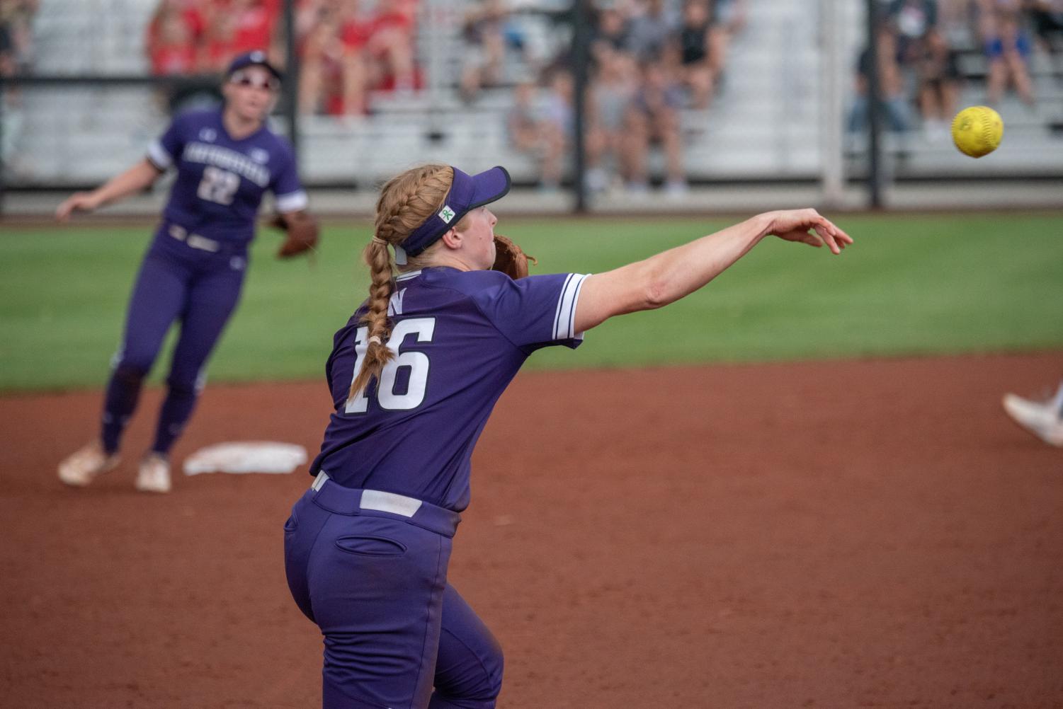  A player wearing a purple jersey on third base throws a softball towards first base.