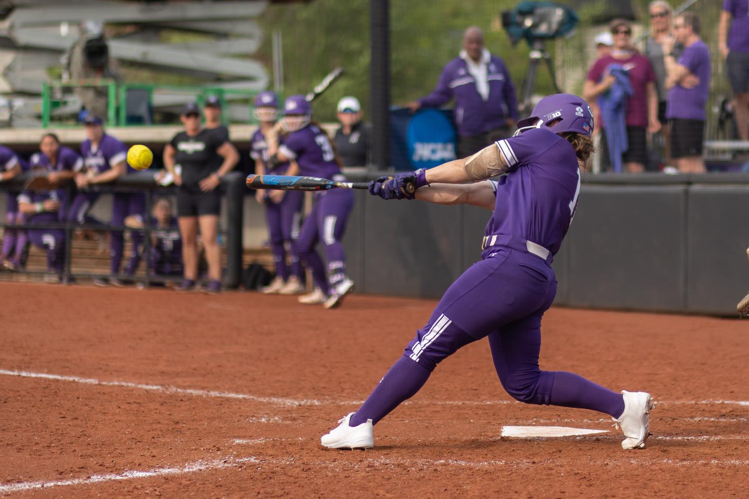 A player in a purple jersey swings and strikes a softball with a bat.