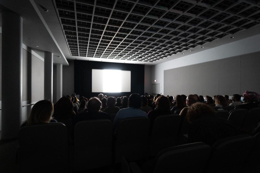 A theater full of people sitting in the dark watch a movie screen at the front of the room.