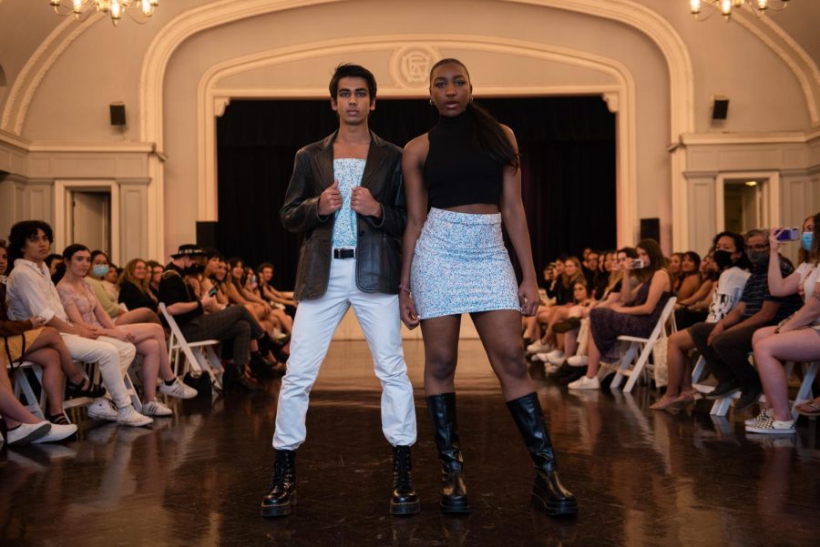 Two models stand in the middle of the frame wearing black, light blue and white while an audience watches them from behind.