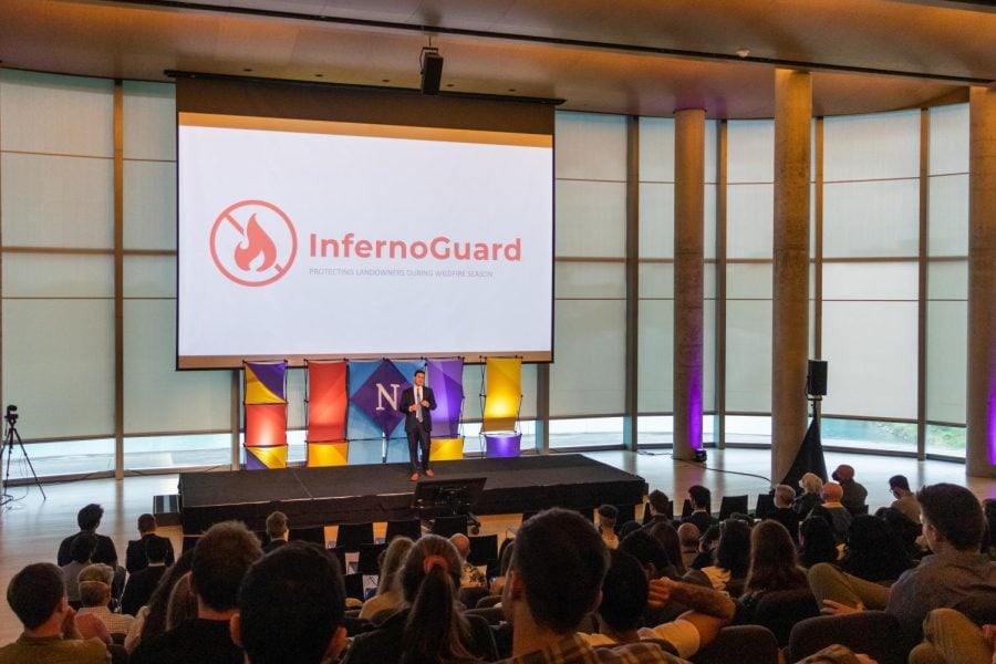 A person stands on a stage in front of a white projector screen that reads “Inferno Guard” in red. An audience sits in chairs in front of the stage.
