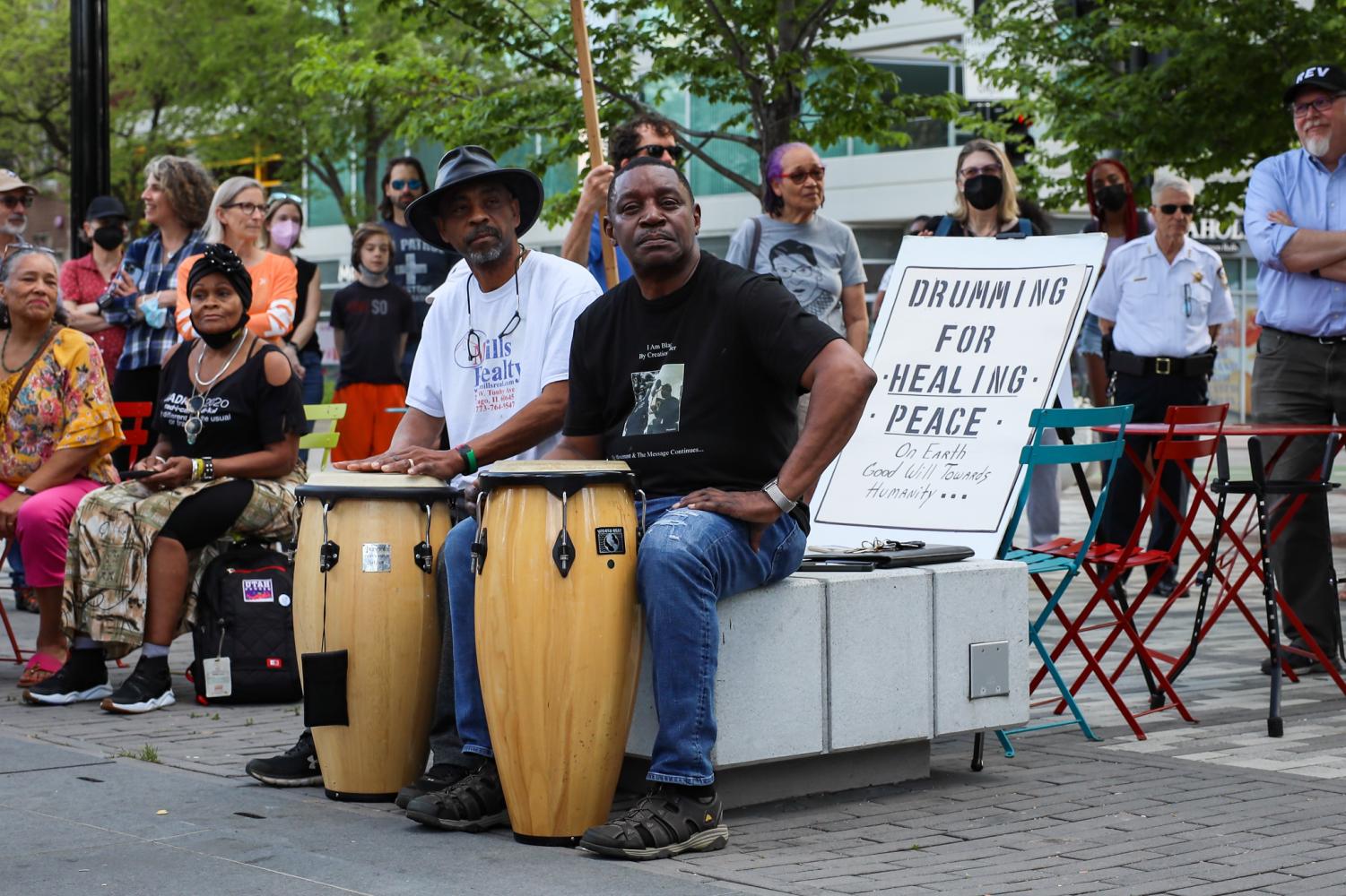 Two people sit behind drums, in front of a sign reading “Drumming for Healing Peace.”