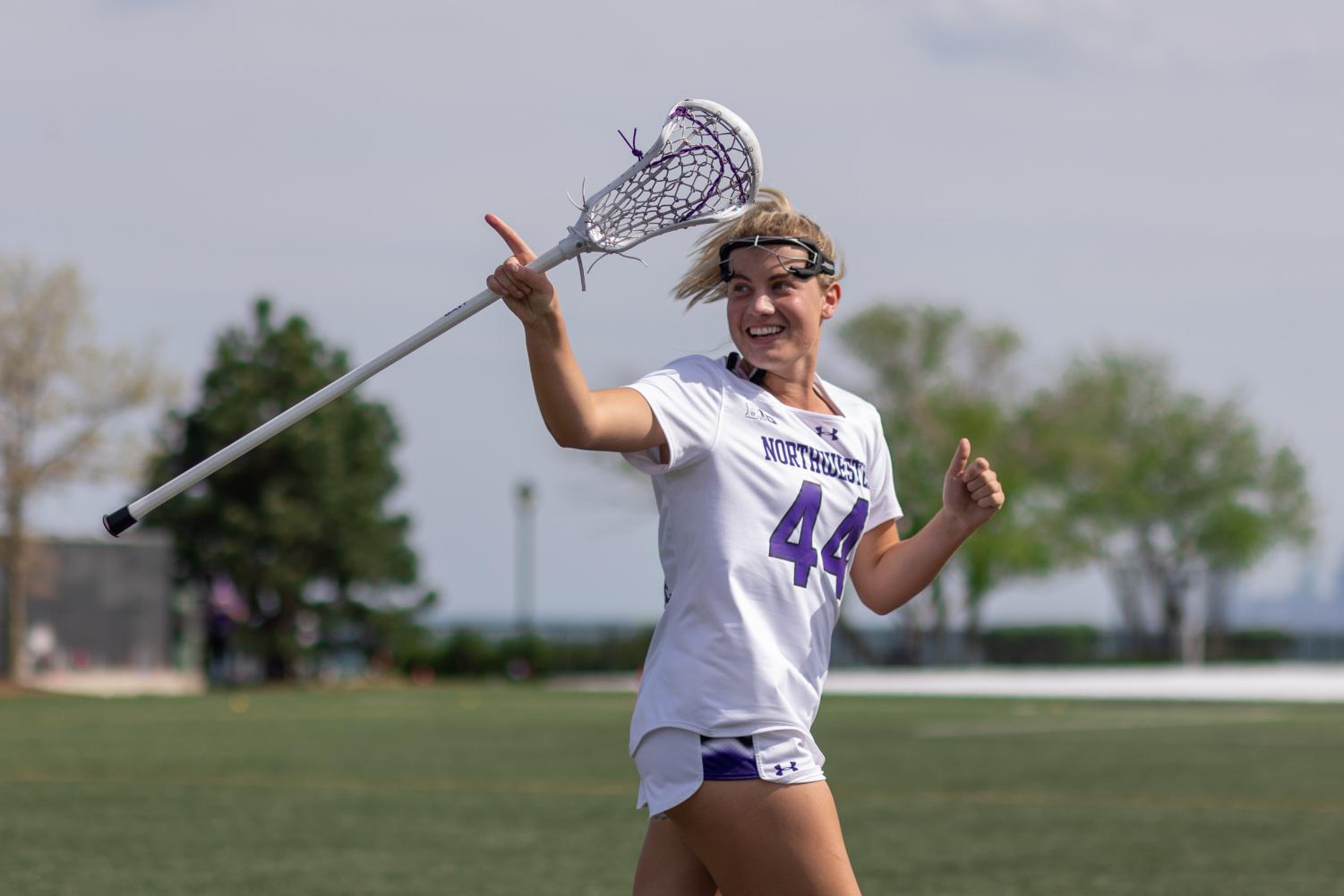 A player in a white jersey holding a lacrosse stick runs and points at their teammates.