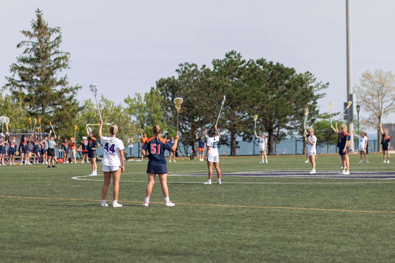 Players in both white and blue jerseys hold up their lacrosse sticks in unison.