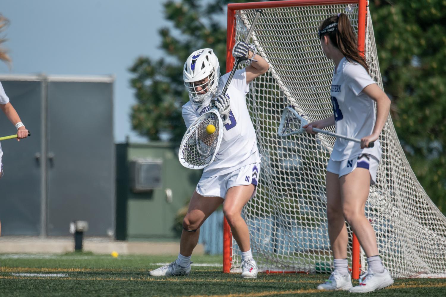 A player in a white jersey holding a goalie lacrosse stick makes a save.