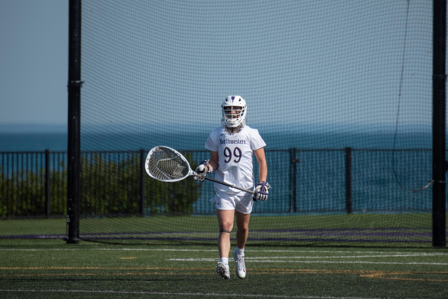 A player wearing a white jersey and holding a goalie lacrosse stick walks across a field.