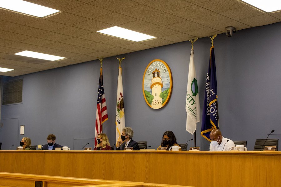 Evanston city councilmembers sit at their desks. Four flags stand behind them.