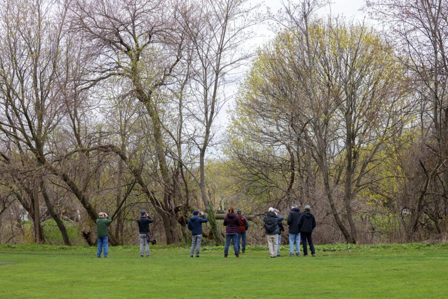Nine individuals with their backs to the camera face a group of trees, many holding binoculars to their faces.