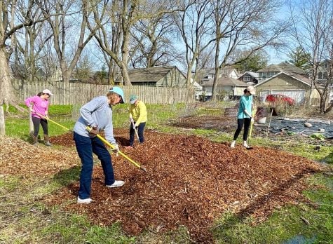 Four gardeners in baseball camps pose holding rakes against a square plot of mulch.