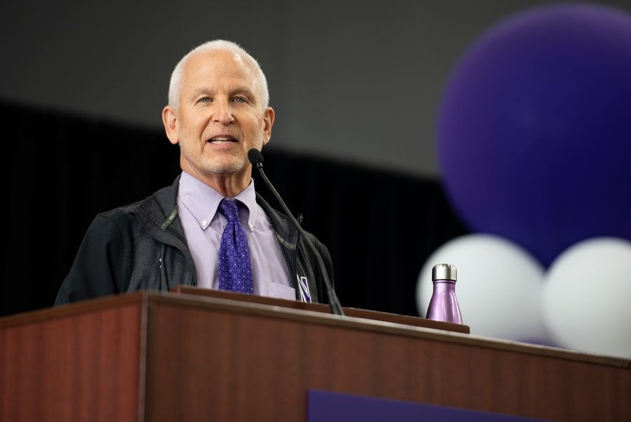 Morton Schapiro stands at a podium wearing a purple shirt and tie, as well as a black windbreaker.