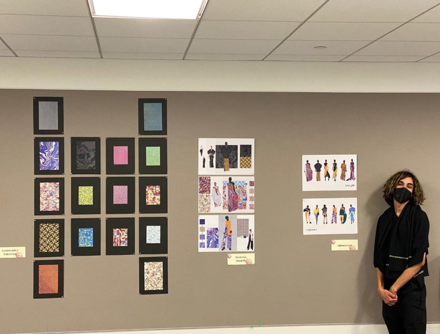 Mayed in an all-black outfit stands in front of a beige wall with prints of their designs