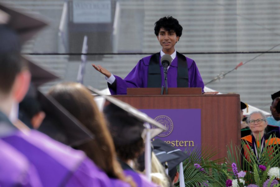 A person speaks at a podium in front of a crowd of people in purple caps and gowns.
