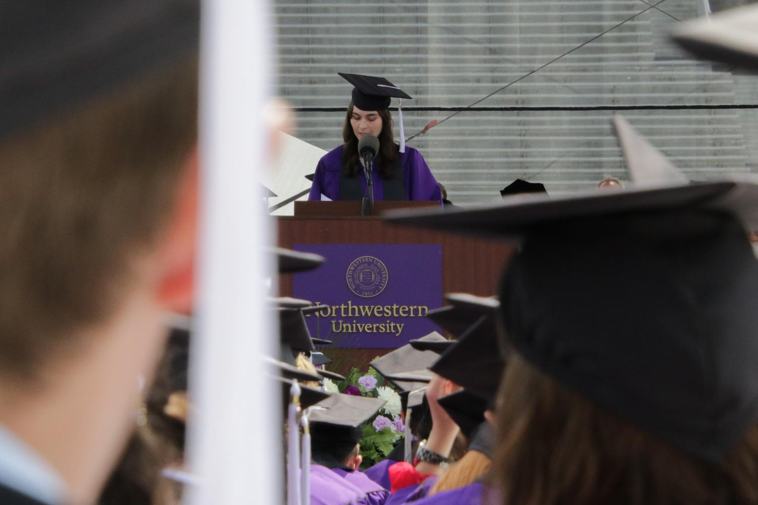 A student in a cap and gown gives a speech in front of a brown podium and a sea of students in similar attire.