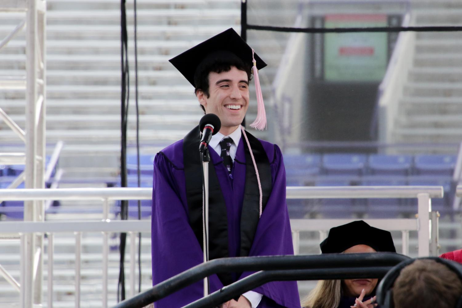 A student in a cap and gown smiles in front of a microphone on stage.