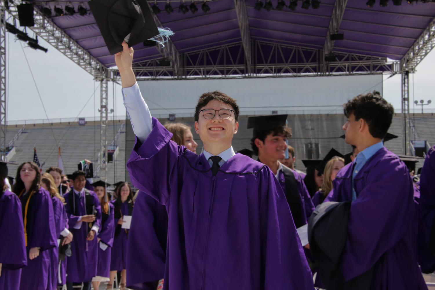 A student in a purple gown raises their arm, with cap in hand, in front of a sea of students.