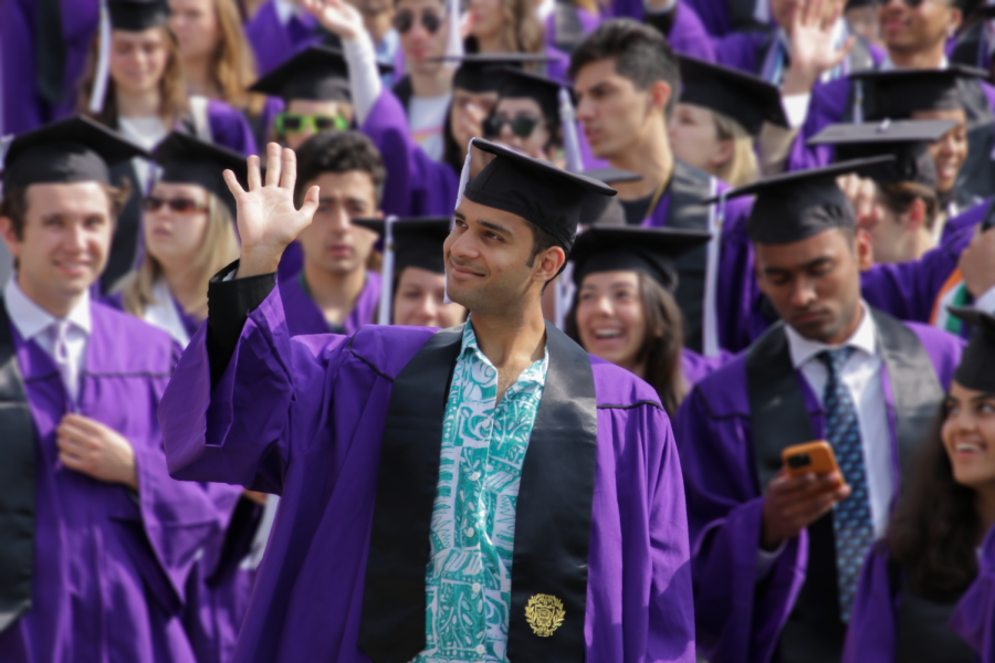 A student in a purple cap and gown waves to their right amid a crowd of purple.