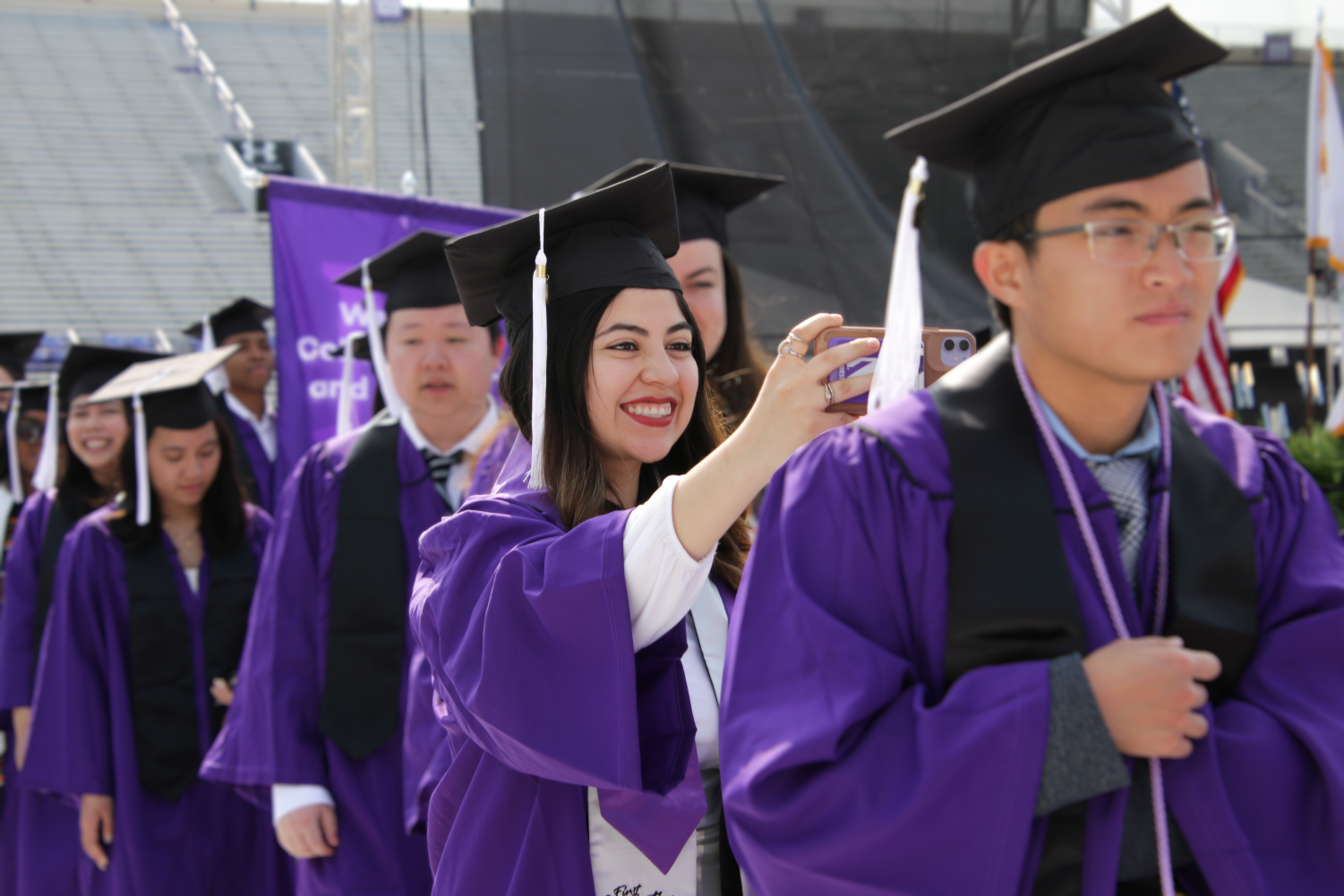 A student looks into their phone camera while walking in a line of people wearing purple gowns.